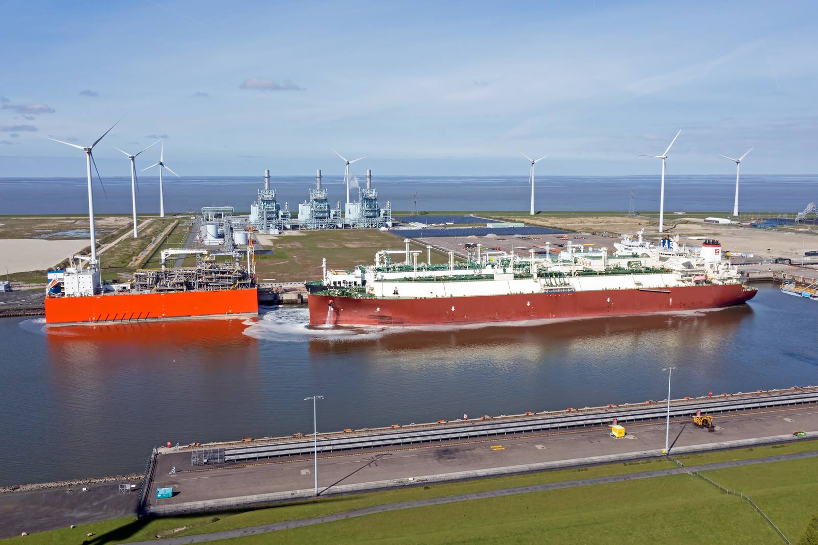 LNG tanker with a red hull next to the red terminal ship in the Eemshaven, with wind turbines and a gas-fired power station in the background