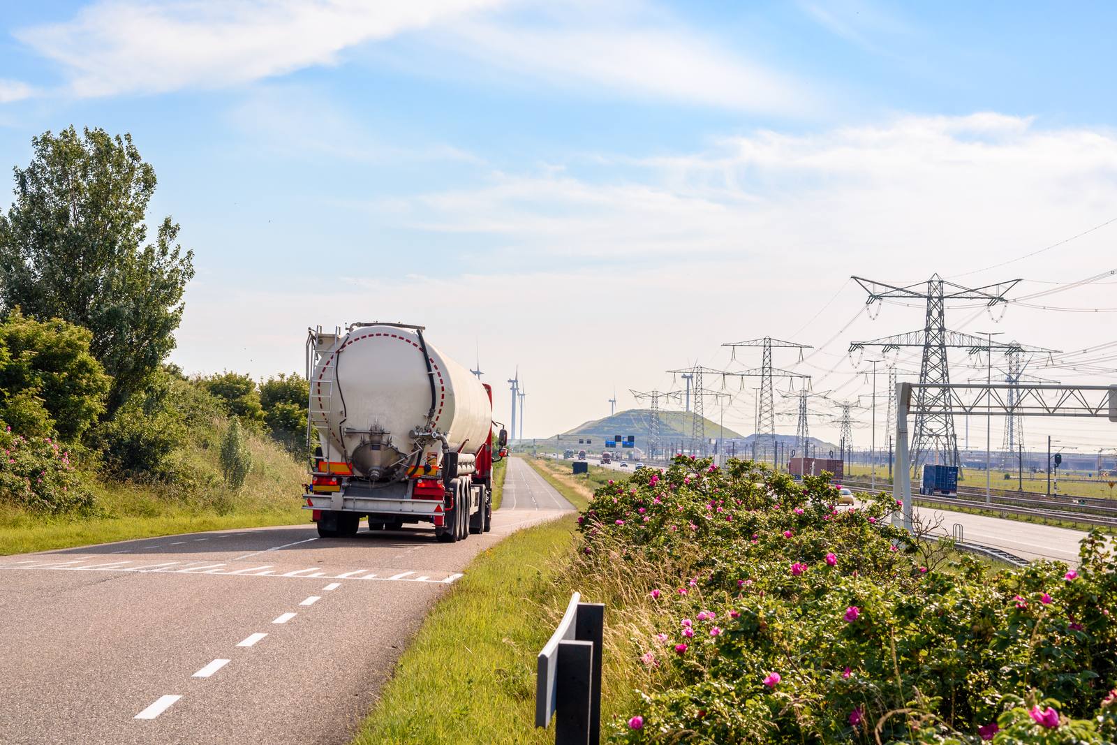 Tanker truck driving on a road in a Dutch port area, with power pylons and wind turbines in the background