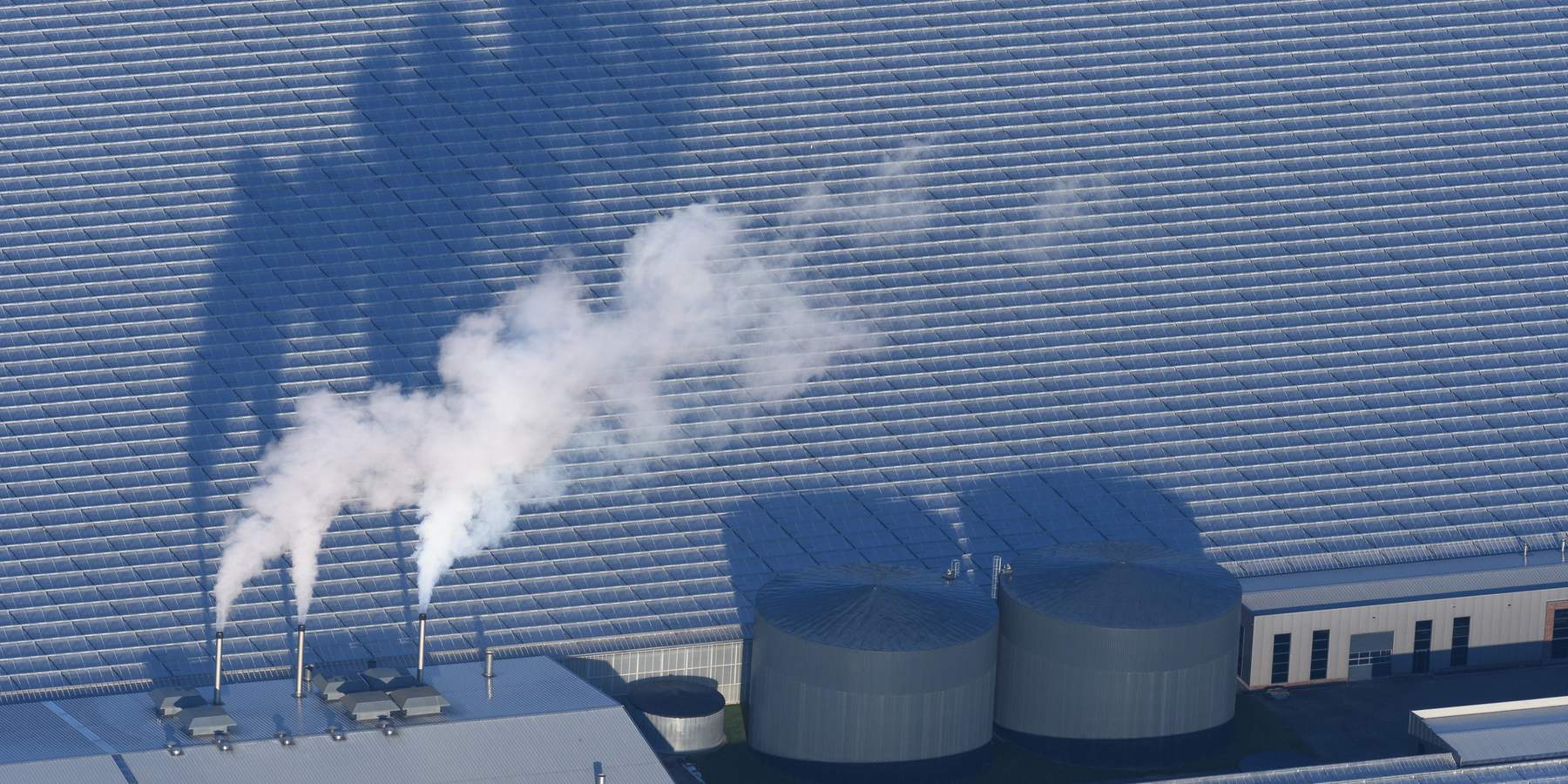 Luchtfoto van een glastuinbouwbedrijf met drie dampende schoorstenen en twee buffertanks